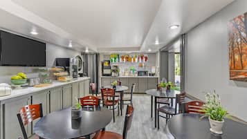 Una cocina moderna con televisor de pantalla plana, refrigerador y microondas. Hay dos mesas de comedor con sillas, y una ventana con vista a árboles.