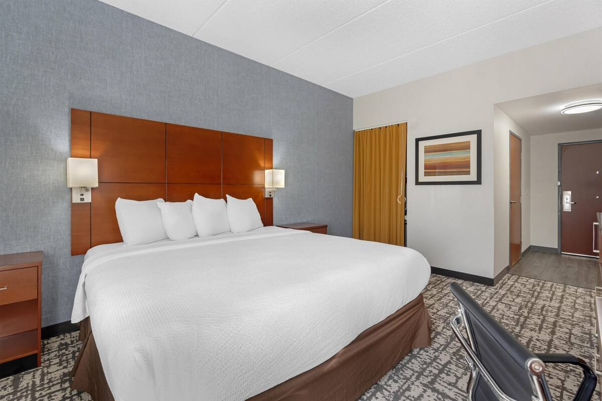 Une chambre d’hôtel avec un grand lit, deux lampes de chevet, une tête de lit, une table de chevet, une chaise, une porte et un tableau encadré au mur.
