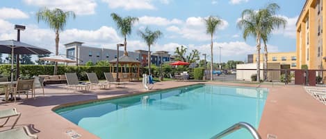 A swimming pool with lounge chairs and umbrellas, surrounded by palm trees and buildings.