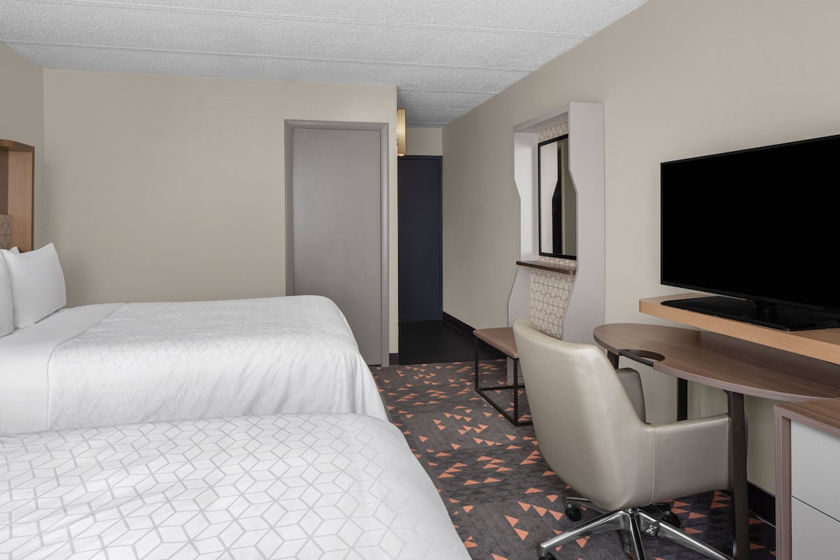 Habitación de hotel con dos camas, un escritorio, una silla, un televisor y una puerta que da a otra habitación.