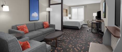 A hotel room with a gray sofa, a small round table, a bed with white linens, a desk with a chair, and a TV.