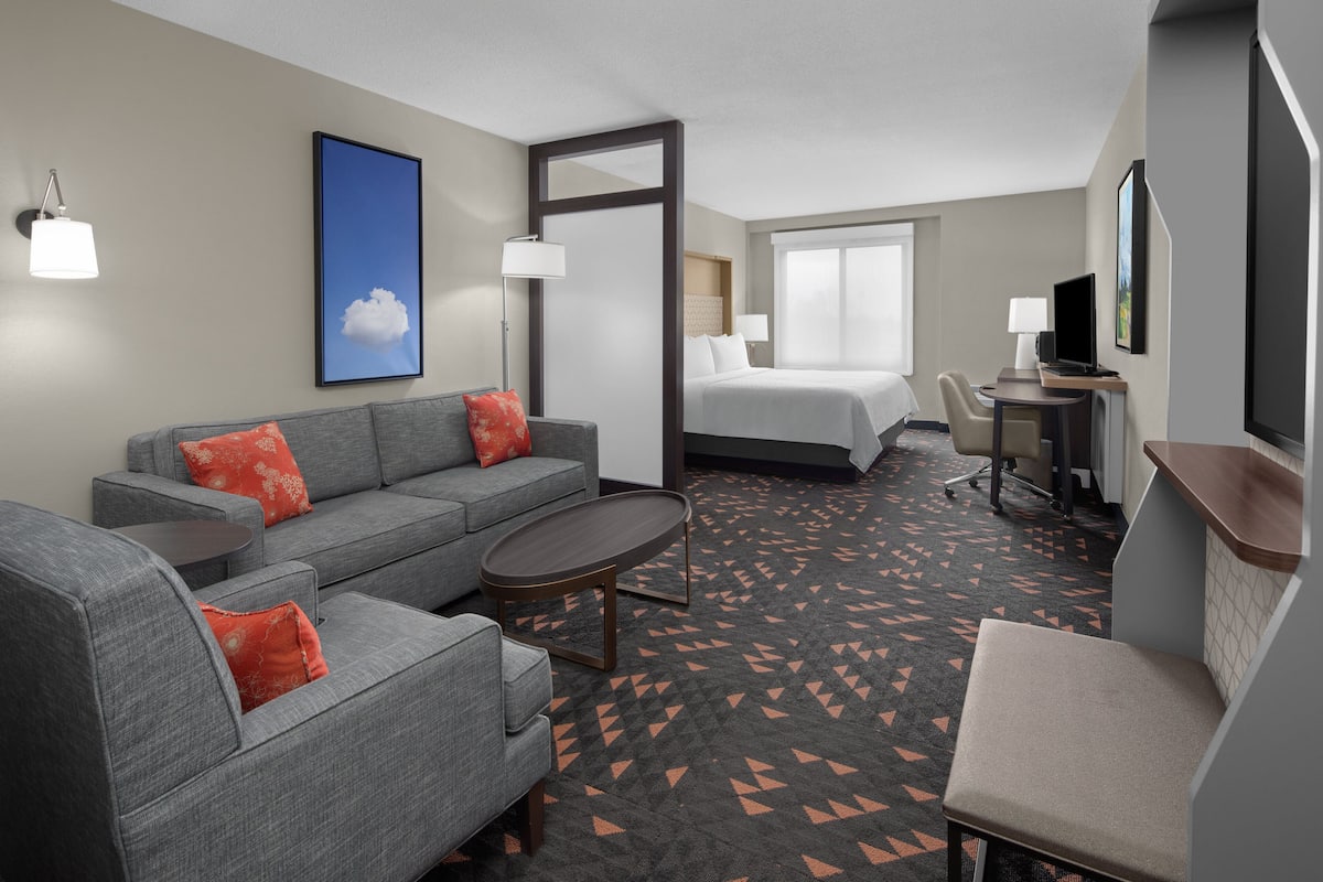 Habitación de hotel con sofá gris, una mesita redonda pequeña, cama, escritorio con silla y televisor.