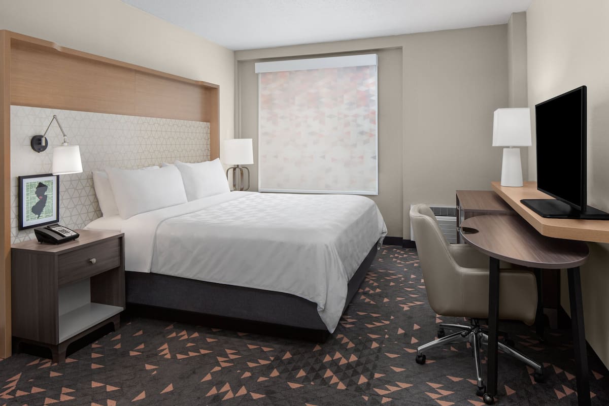 Habitación de hotel con cama, mesitas de noche, escritorio, silla, televisor y ventana con persianas.