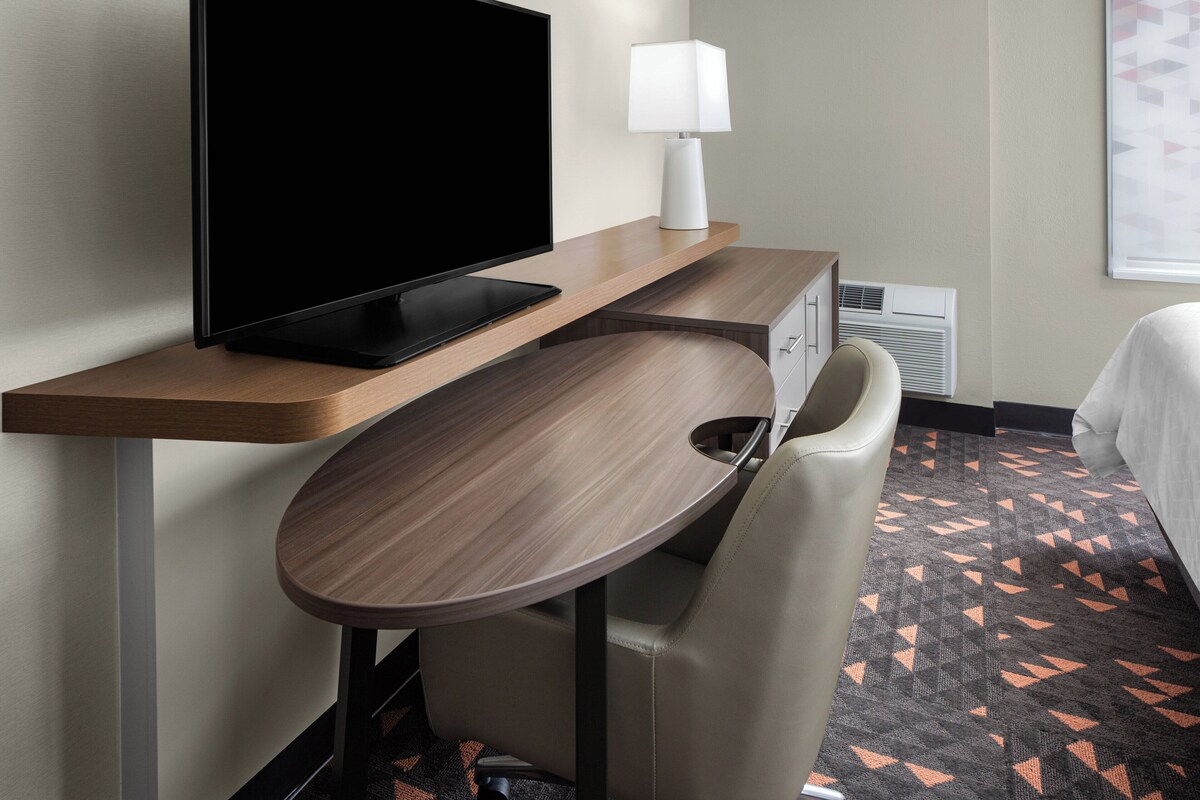 Une chambre d’hôtel avec un téléviseur à écran plat posé sur un bureau en bois, une lampe blanche, un fauteuil beige et une moquette à motifs.