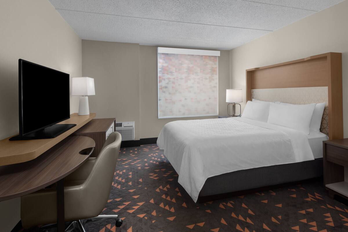 Habitación de hotel con cama, escritorio con televisor, silla, ventana con persiana estampada y alfombra de diseño geométrico.