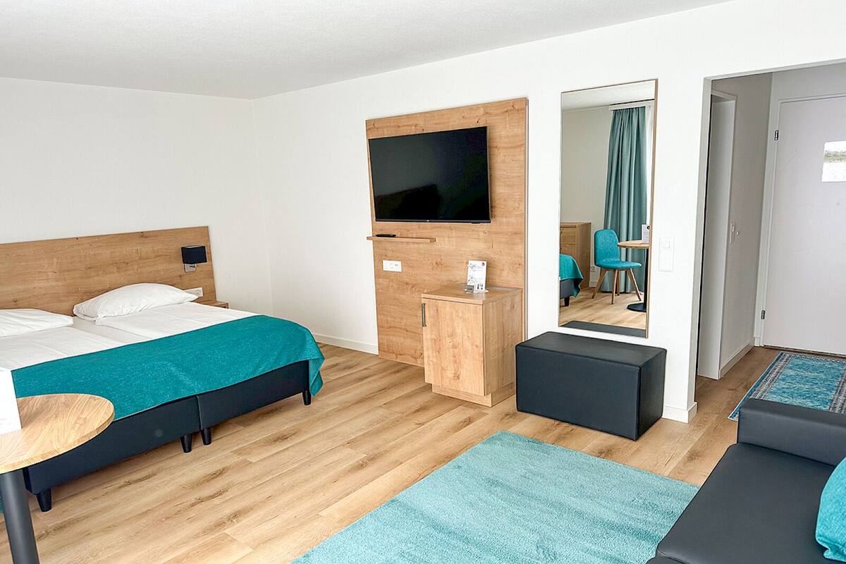 A modern hotel room with a bed, a desk, a TV, a mirror, and a seating area.