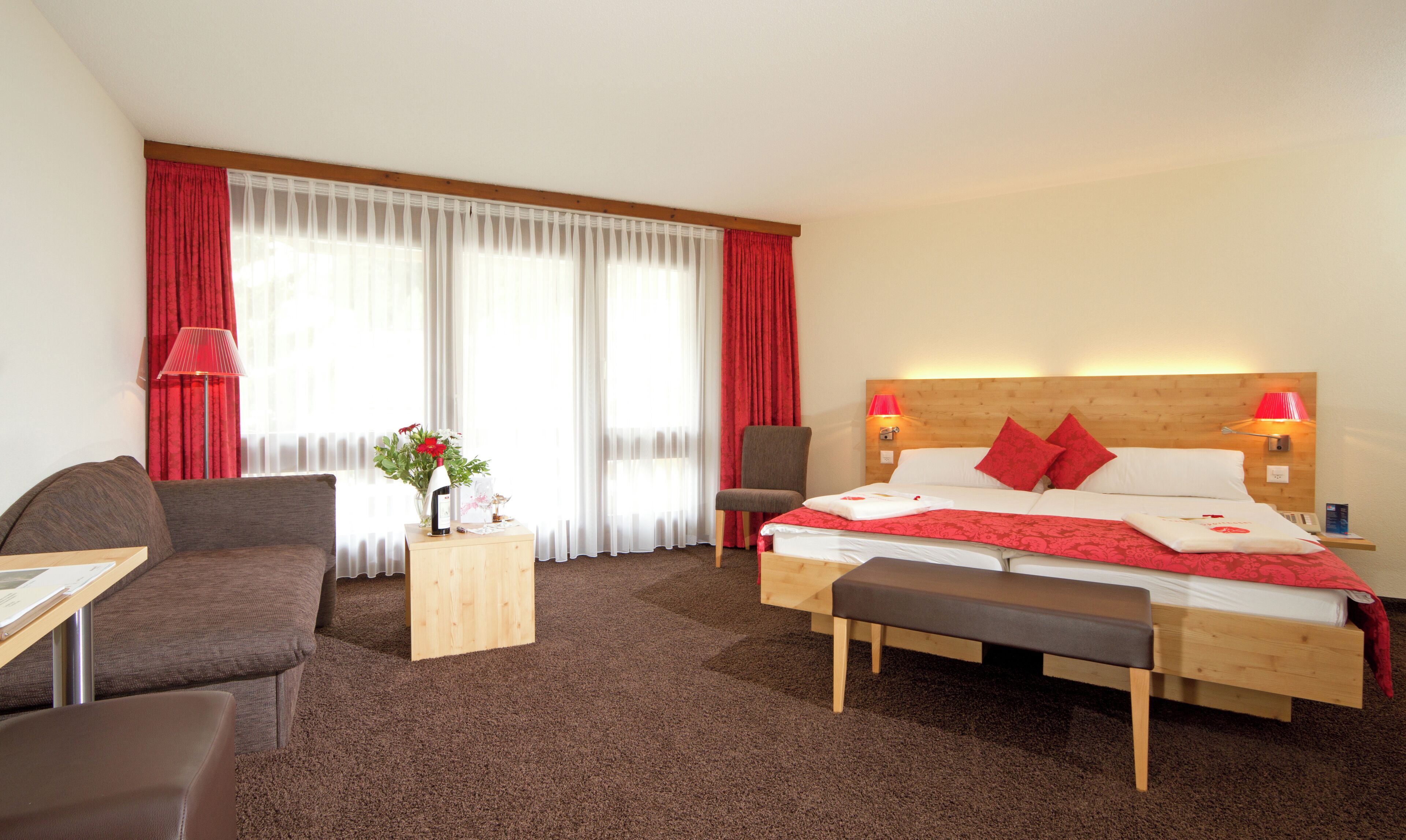 A hotel room with a bed, two chairs, a small wooden table, a lamp, and a window with red curtains.