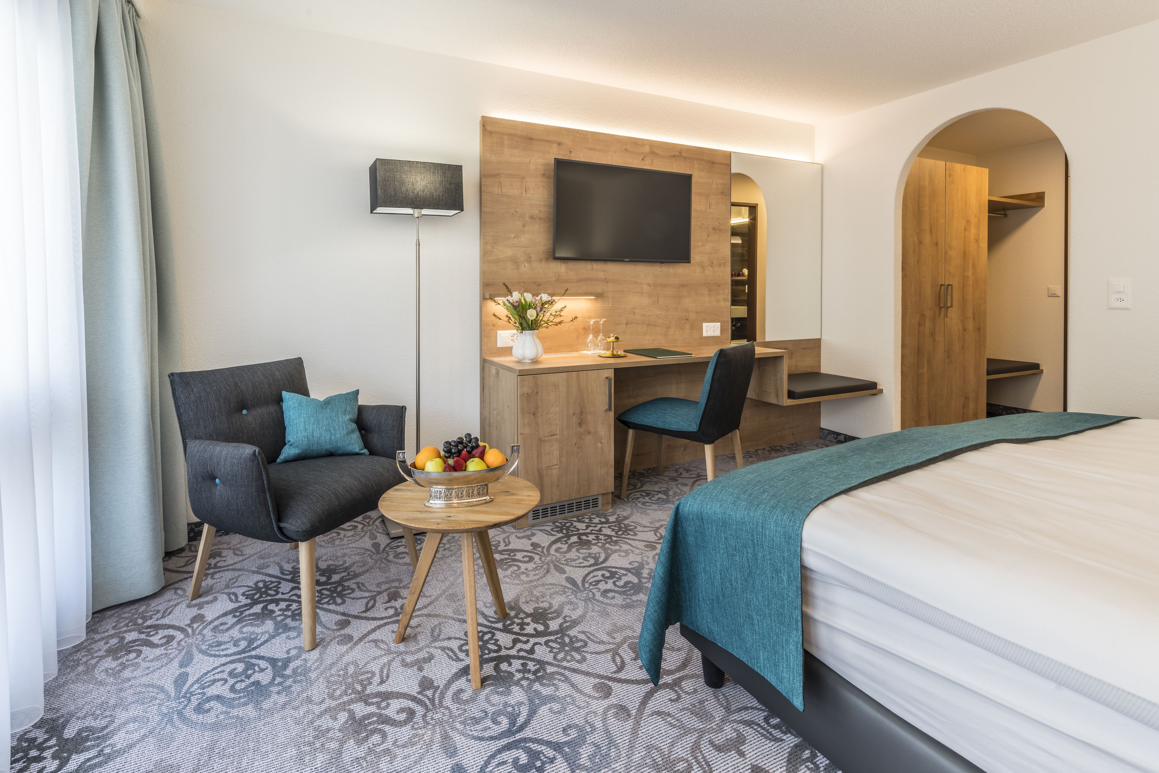 A hotel room with a bed, a desk with a chair, a small table with a fruit bowl, a TV mounted on the wall, and a wooden wardrobe.