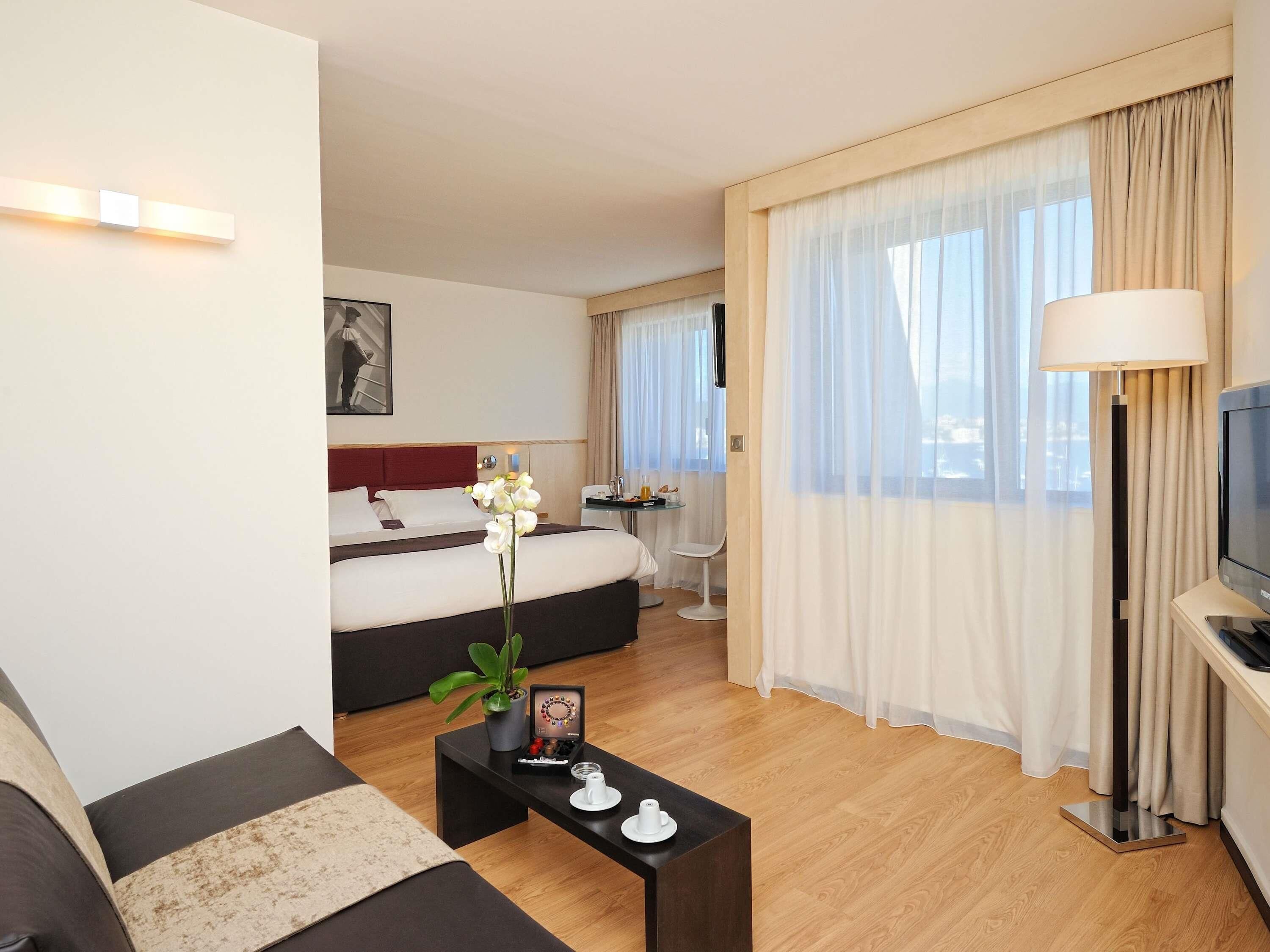 Una habitación de hotel moderna con una cama, un sofá, una mesa de centro con una planta y un marco de fotos, un televisor y una zona de comedor con mesa y sillas.