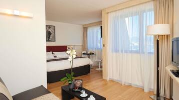 A modern hotel room with a bed, a sofa, a coffee table with a plant and a picture frame, a TV, and a dining area with a table and chairs.