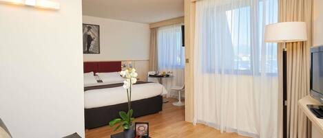 Una habitación de hotel moderna con una cama, un sofá, una mesa de centro con una planta y un marco de fotos, un televisor y una zona de comedor con mesa y sillas.