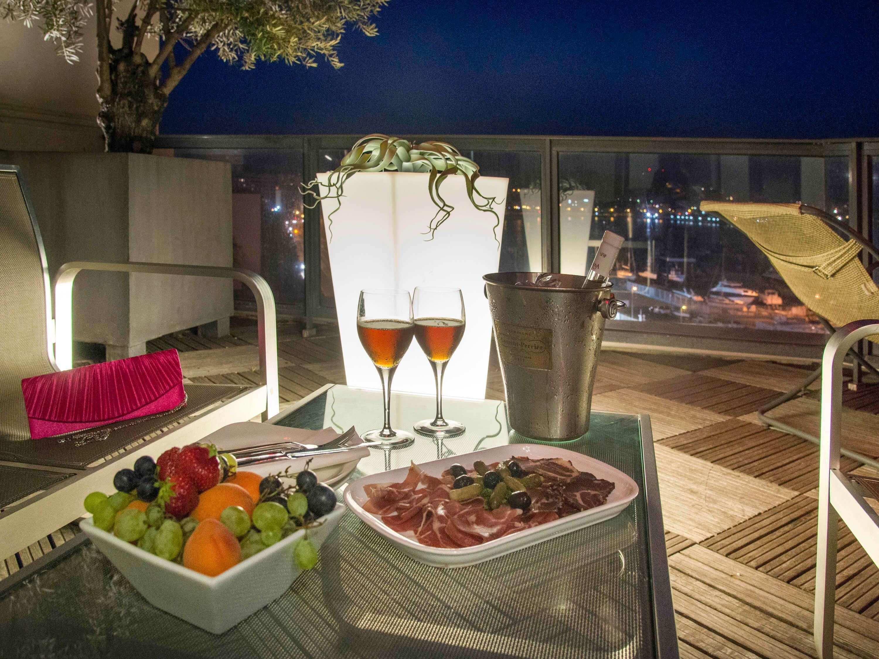 A table with a fruit bowl, a plate of meat, two glasses of wine, and a metal bucket on a balcony at night.