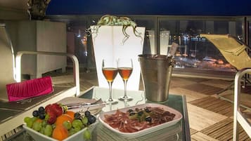 A table with a fruit bowl, a plate of meat, two glasses of wine, and a metal bucket on a balcony at night.