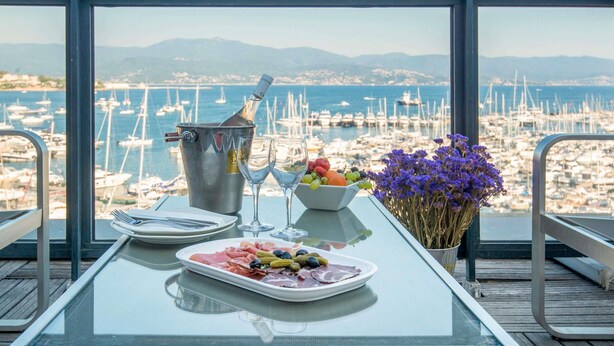 Una mesa con cubierta de vidrio, una botella de champán, un cuenco de fruta y un plato de comida, con vista a un puerto deportivo lleno de barcos.
