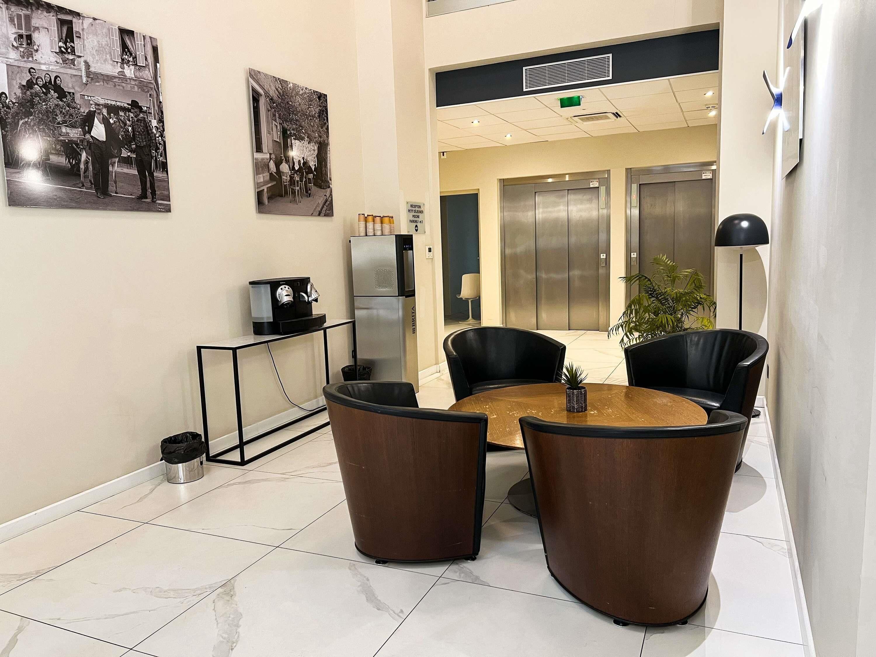 Un hall d’entrée avec une machine à café, deux fauteuils noirs, une table ronde et une plante.