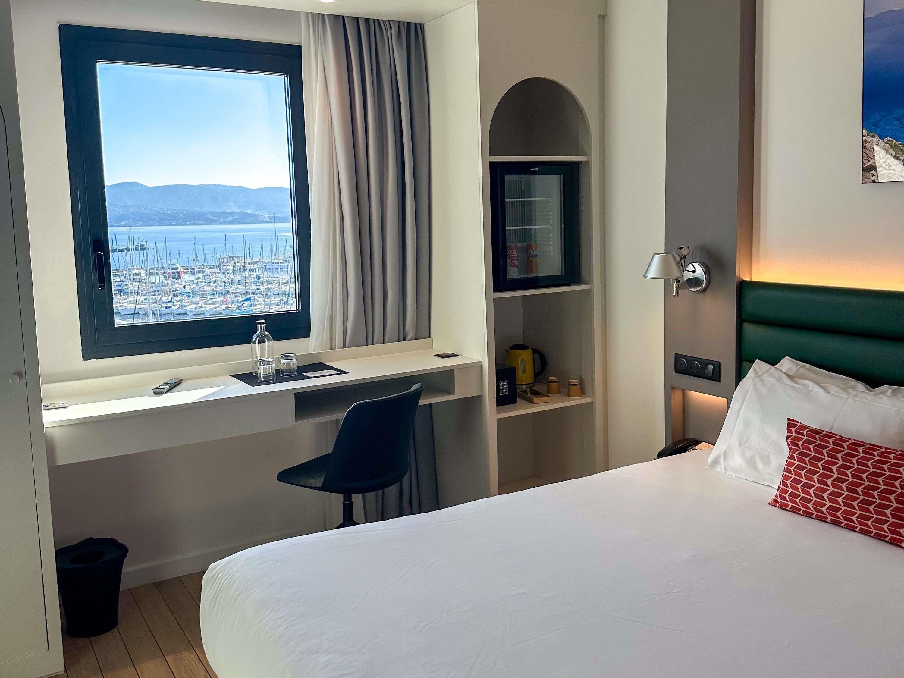 A hotel room with a bed, a desk with a computer, a chair, a window with a view of a marina, and a shelf with amenities.