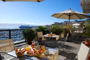 Una terraza con una mesa dispuesta para el desayuno, un ordenador portátil y vistas al puerto deportivo y al paisaje urbano.