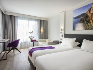 A hotel room with a bed, a desk, a chair, a small table with a plant, and a painting of a coastal landscape. - Mercure Ajaccio Centre (Ajaccio)