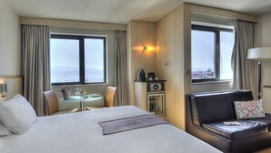 Habitación de hotel con una cama grande, zona de comedor, sofá, minibar y vista al mar.