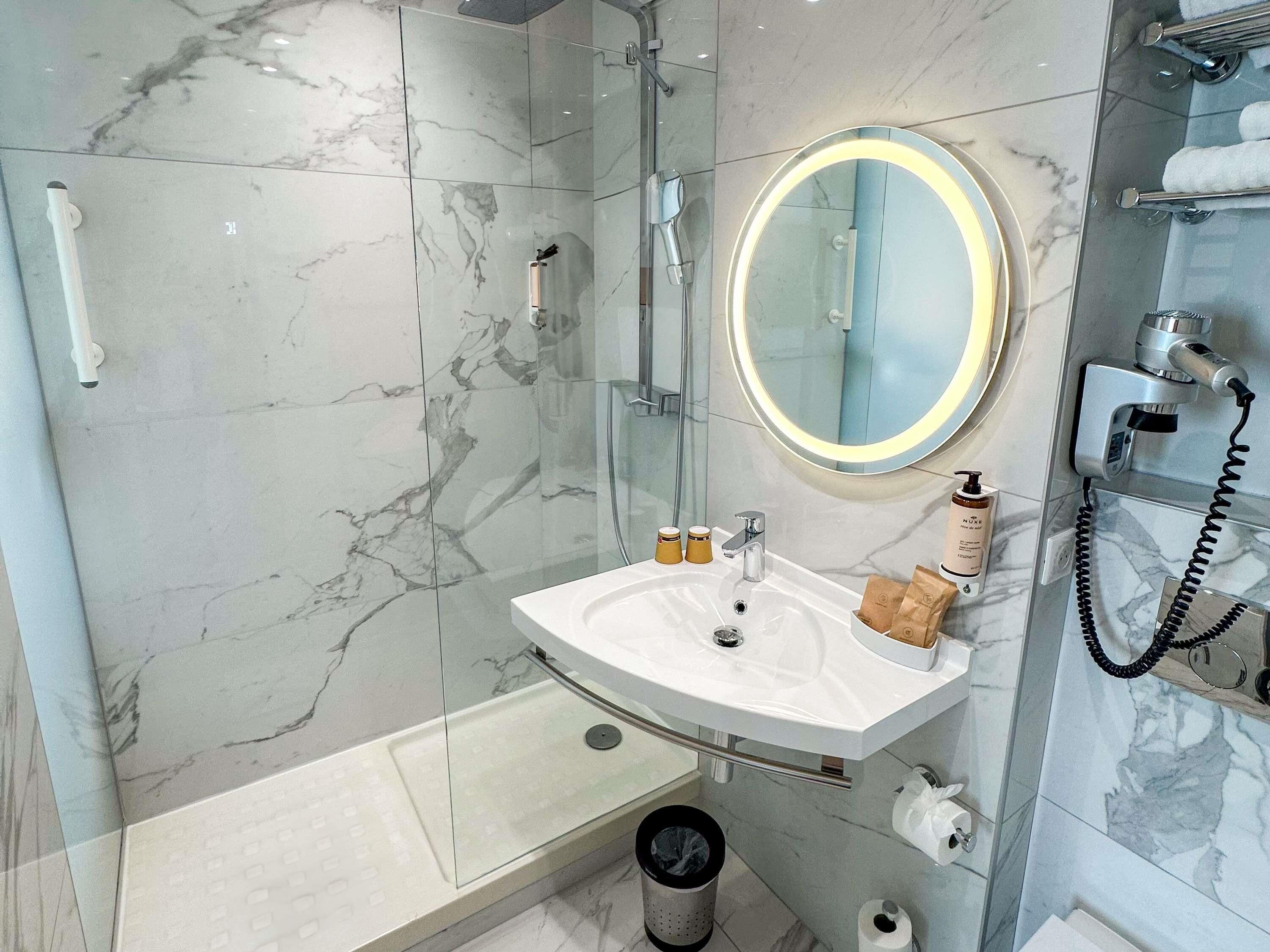 Un baño moderno con ducha de vidrio, lavamanos blanco y espejo, además de artículos de aseo.