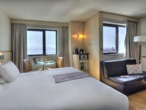 A hotel room with a large bed, a dining area with a table and chairs, a sofa, a minibar, and a view of the sea.