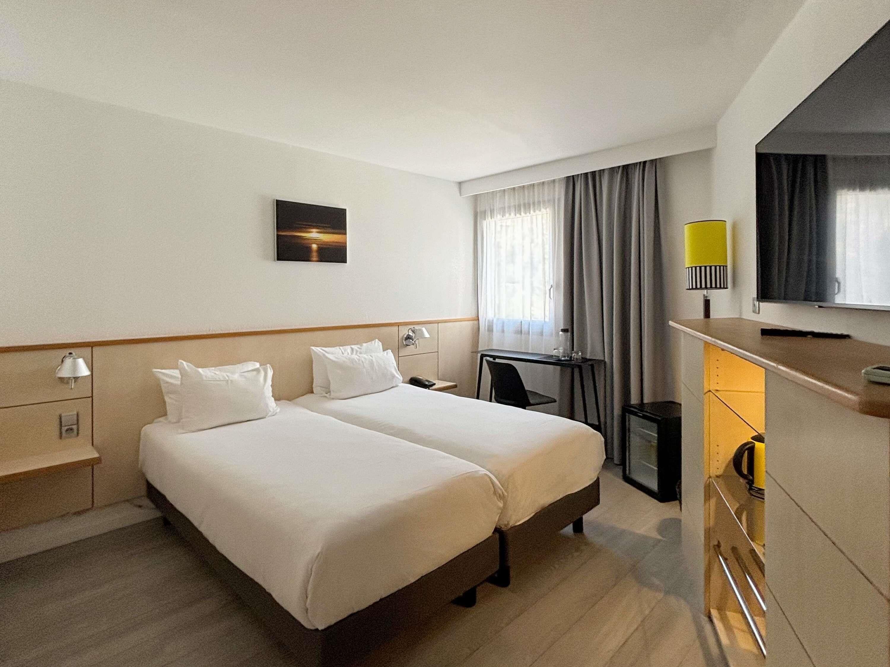 Ein Hotelzimmer mit zwei Betten, einem Schreibtisch, einem Fernseher und einem Vorhangfenster.