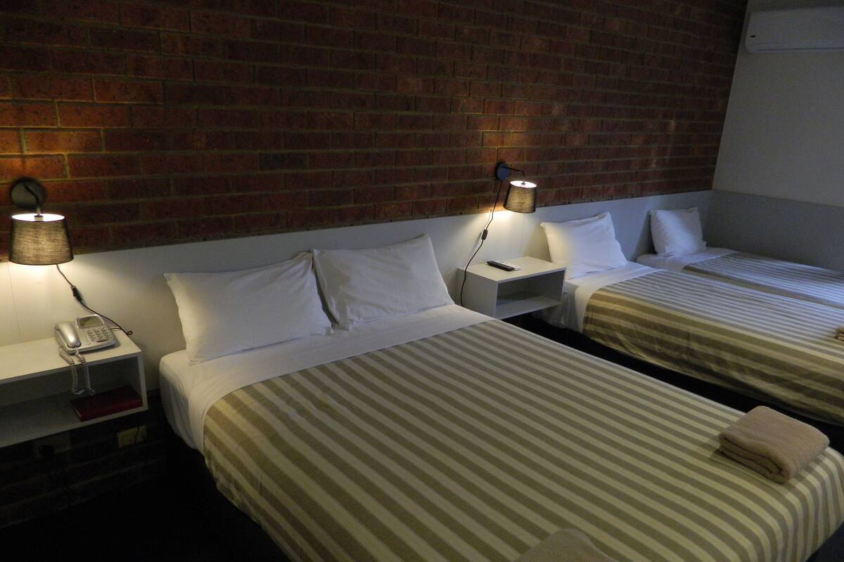 A hotel room with two beds, each with striped bedding and white pillows. There are bedside lamps, a phone on the nightstand, and a brick wall behind the beds.