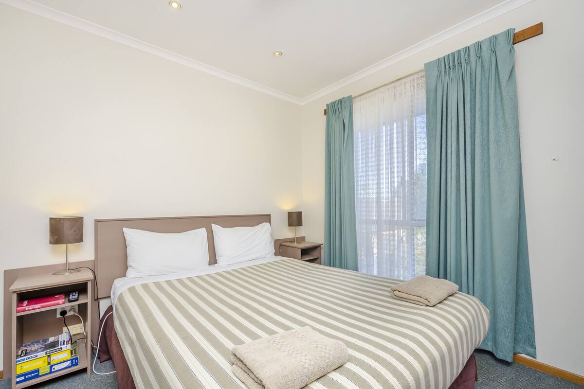A hotel room with a large bed, two bedside tables, a nightstand, a lamp, and a window with curtains.