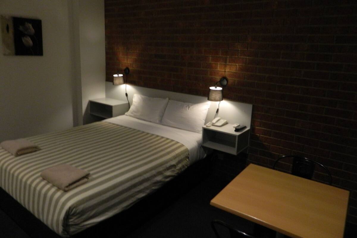 A hotel room with a bed, two bedside tables, a nightstand, a small table, and a brick wall.