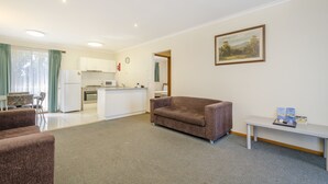 A living room with a brown sofa, a white table with brochures, a kitchen in the background, and a painting on the wall. - Werribee Motel & Apartments (Werribee)