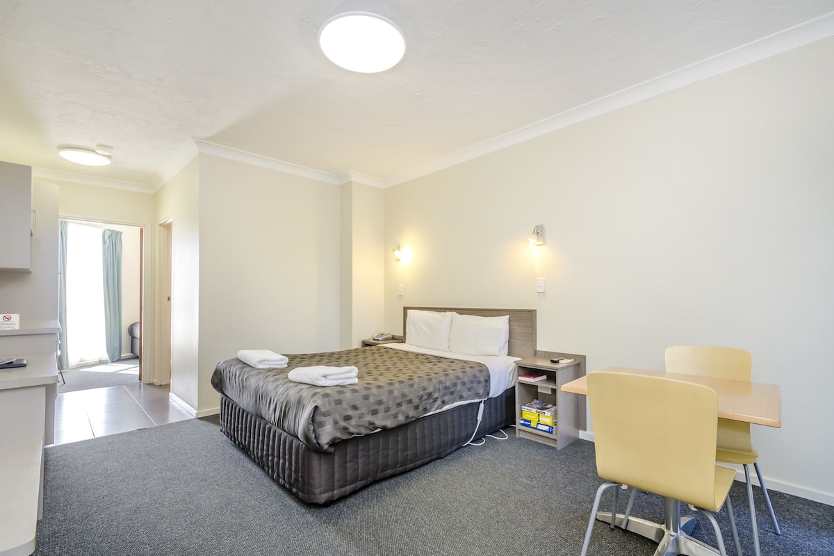 A hotel room with a bed, bedside table, desk, chair, and a small kitchenette area.