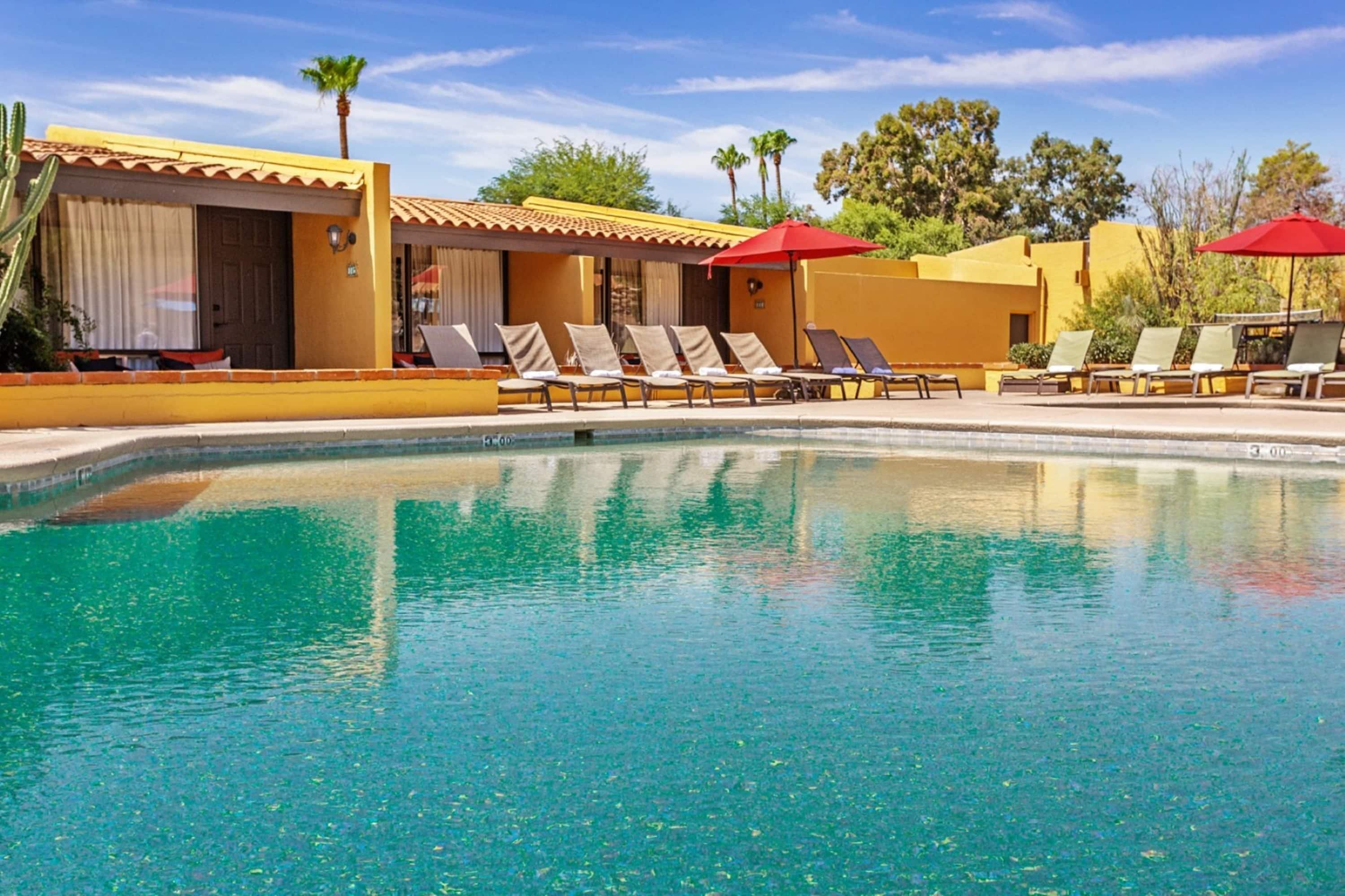 A pool area with lounge chairs and umbrellas, a building with yellow walls and a tiled roof, and palm trees in the background.