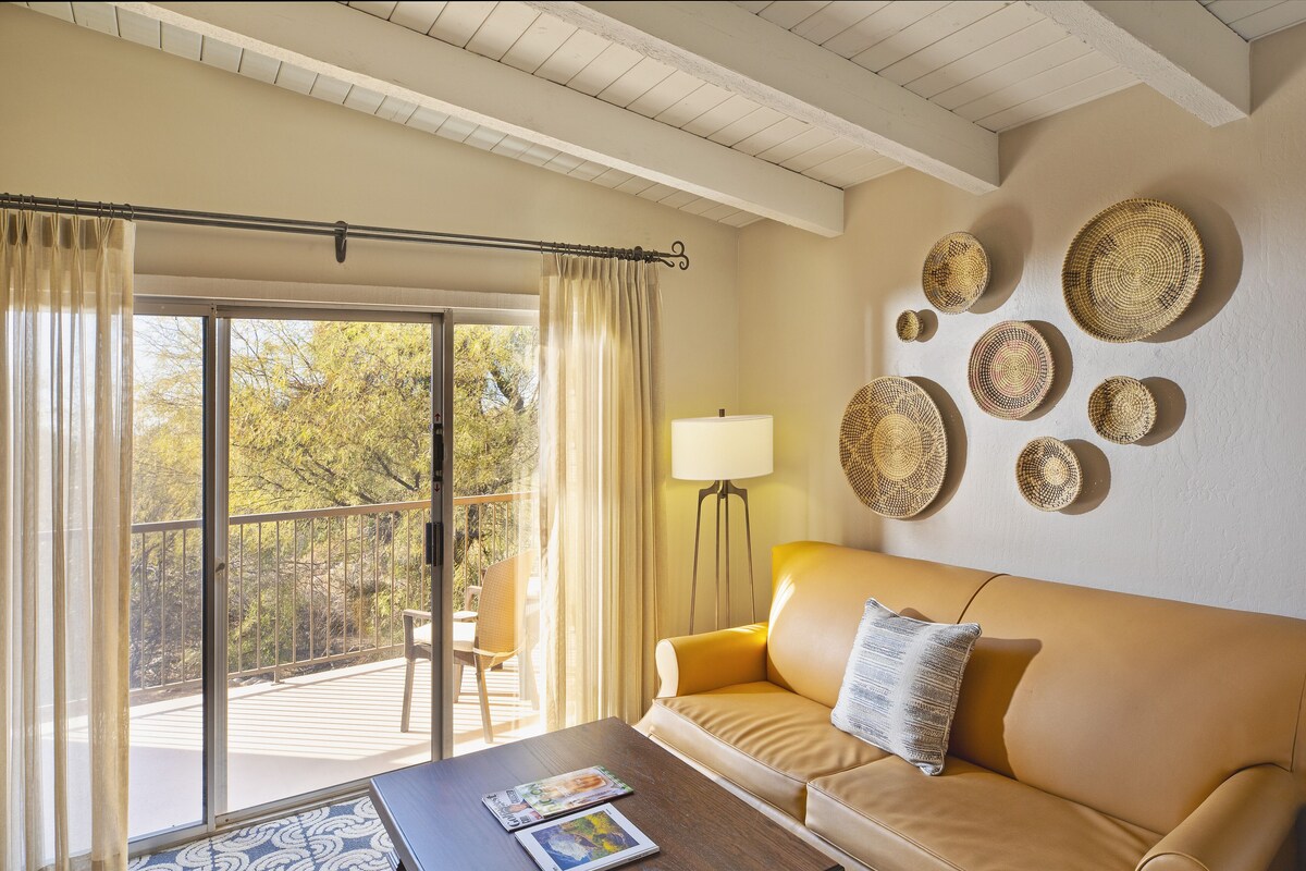 A living room with a sofa, a coffee table with magazines, a floor lamp, and a view of trees through a sliding door.