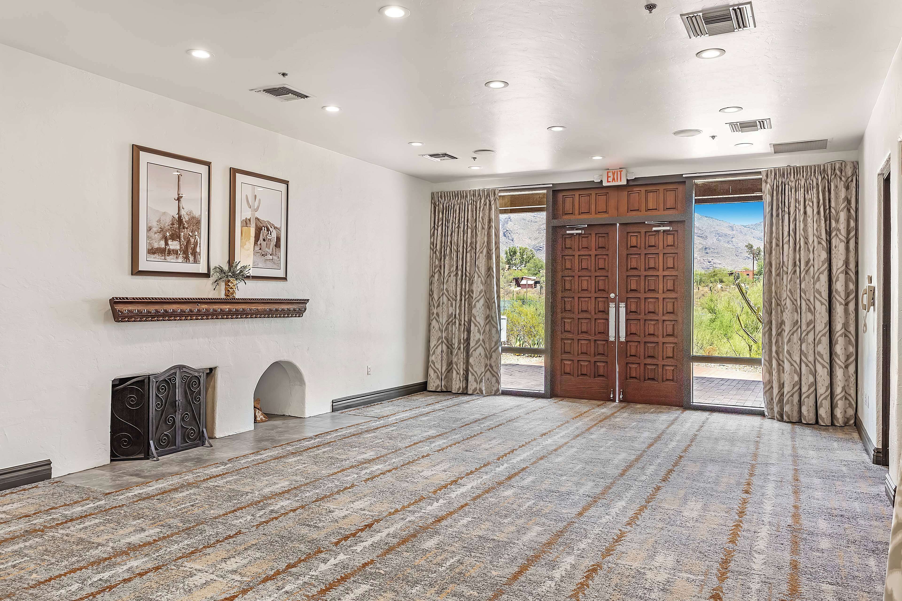 A spacious room with a patterned carpet, a fireplace, framed pictures, and a large wooden door leading to a scenic outdoor view.