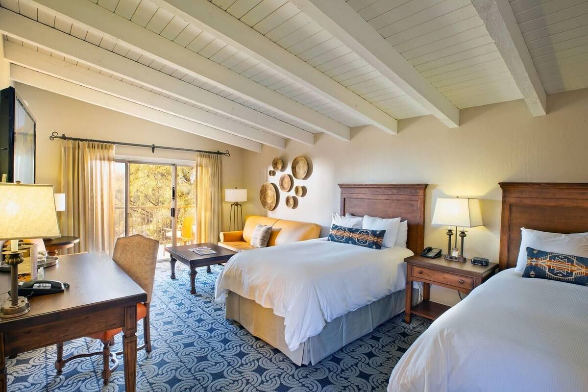 A hotel room with two beds, a desk, a chair, a small table, and a nightstand with a lamp.