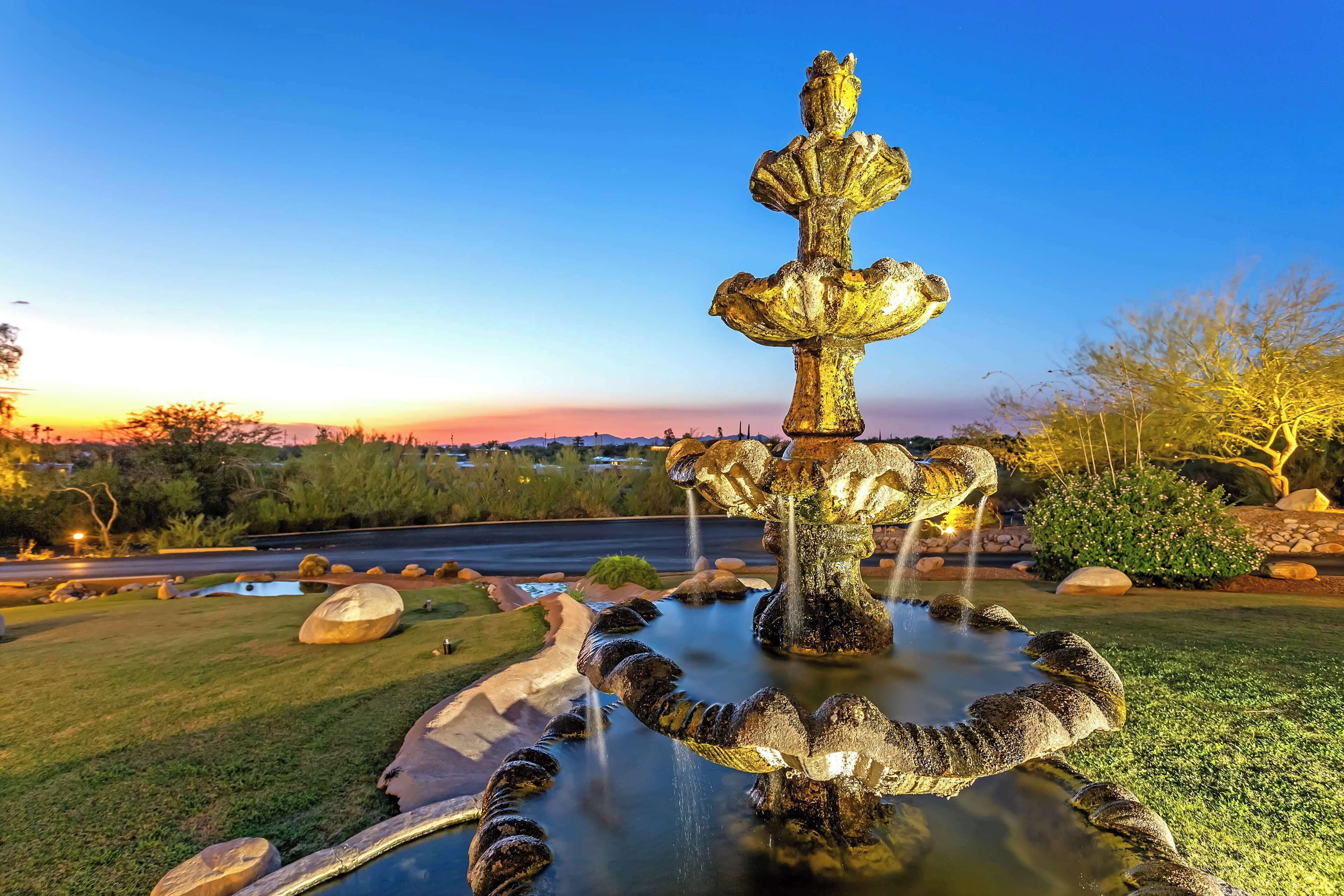 A fountain with multiple tiers and water spouts, surrounded by rocks and grass, with a clear sky in the background.