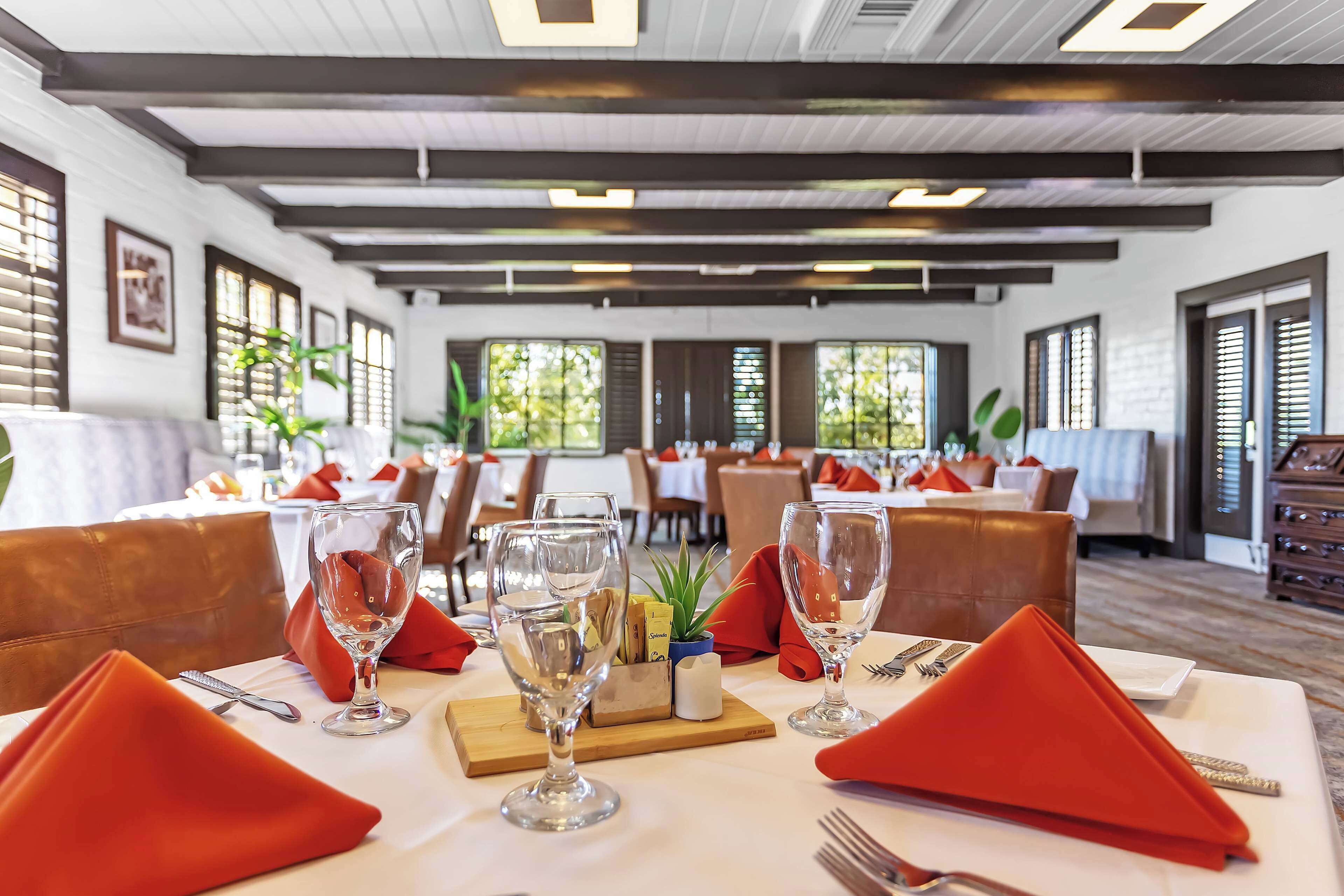 A restaurant with tables set for dining, featuring red napkins, wine glasses, and a wooden board with a plant.