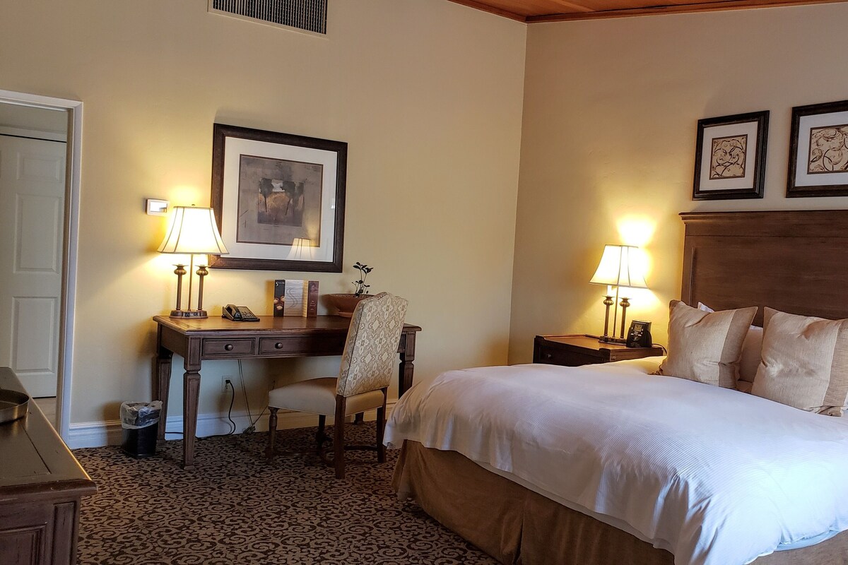 A hotel room with a large bed, two bedside tables with lamps, a desk with a chair, a framed picture on the wall, and a patterned carpet.