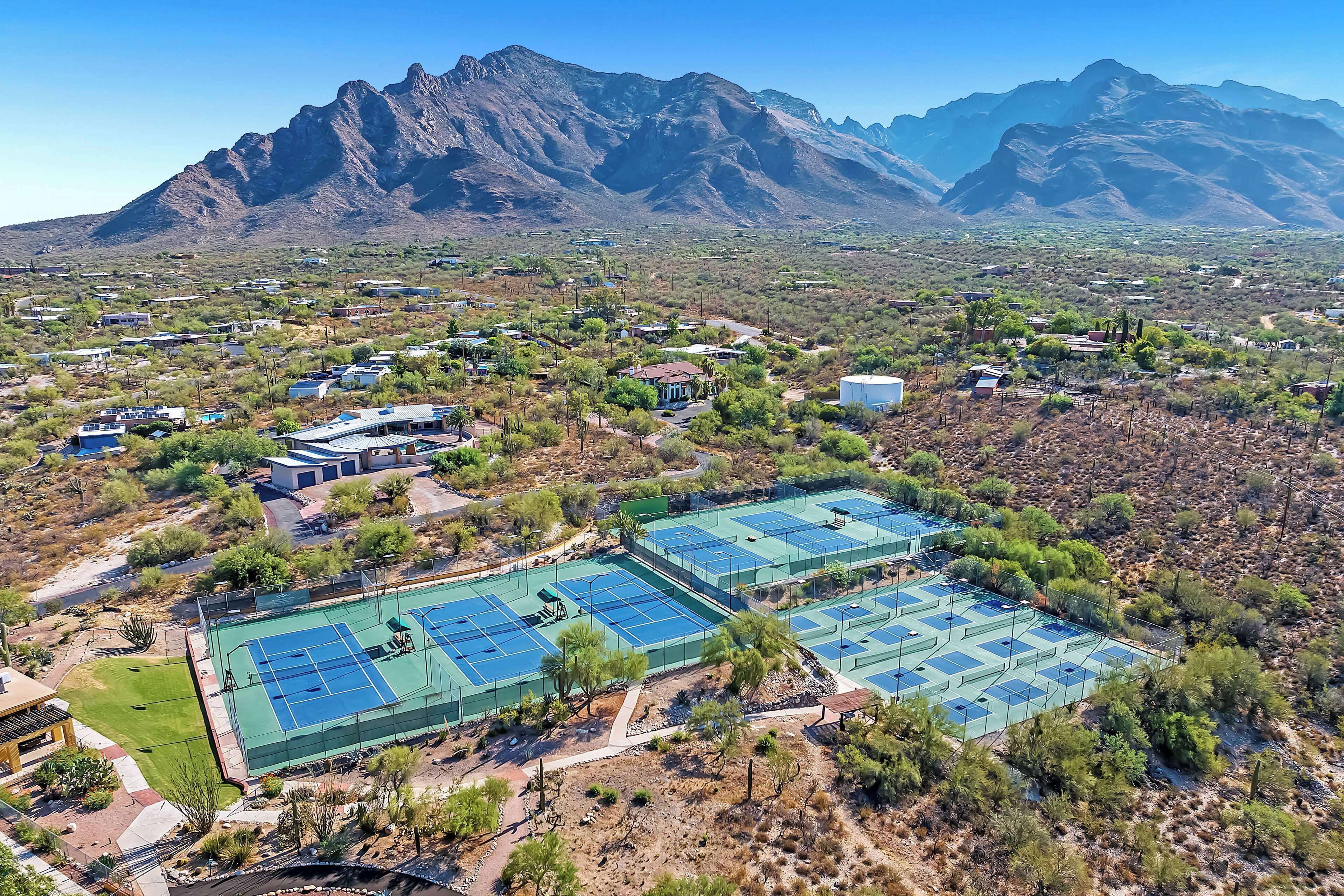 An aerial view of a residential area with multiple tennis courts surrounded by sparse vegetation and houses.