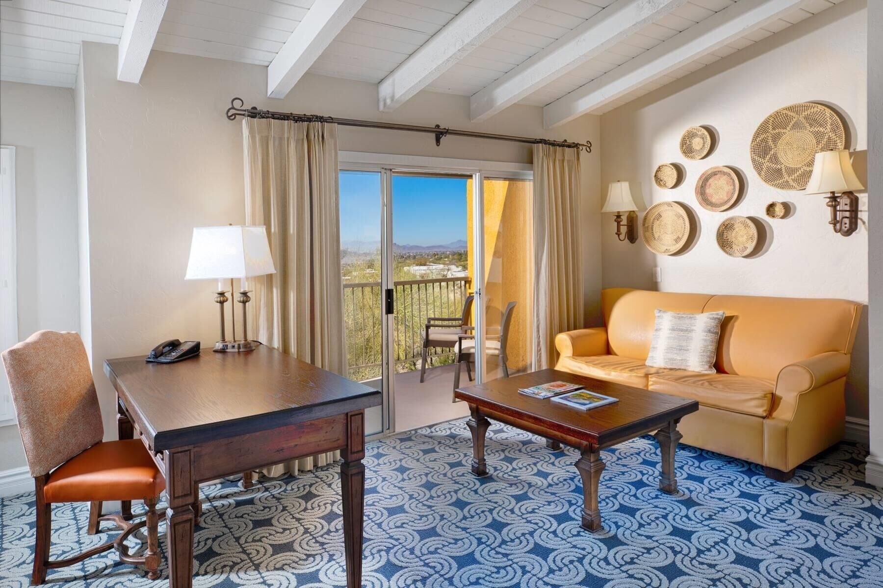 A room with a wooden table, a sofa, a patterned carpet, and a balcony with a view.