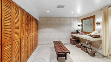 Baño con taquillas de madera, un banco, lavamanos con artículos de aseo y un espejo.