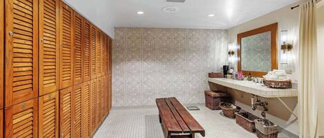 A bathroom with wooden lockers, a bench, a sink with toiletries, and a mirror.