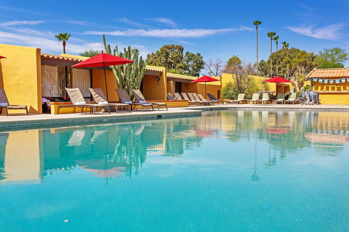 A pool area with sun loungers and umbrellas, surrounded by yellow buildings and palm trees.