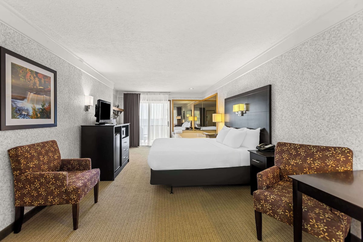 A hotel room with a bed, two armchairs, a desk, a TV, and a mirror.