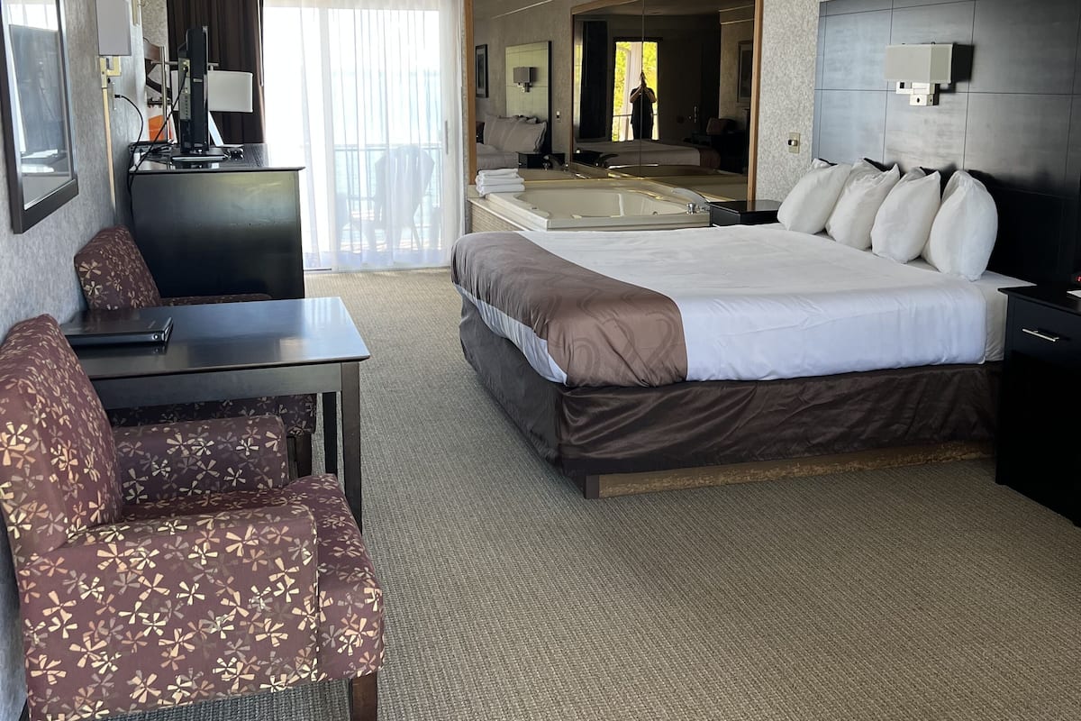 A hotel room with a large bed, a desk, a chair, a TV, and a bathroom visible through a sliding door.