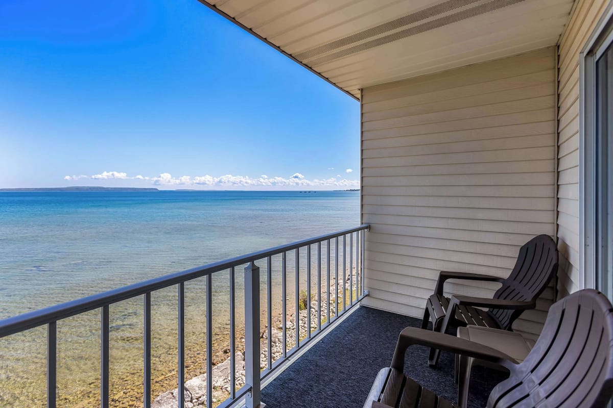 A balcony with a clear view of the ocean, two Adirondack chairs, and a metal railing.