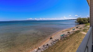 Una vista despejada de una playa con costa rocosa, algunas sillas de descanso y una línea de costa distante con árboles bajo un cielo azul claro.