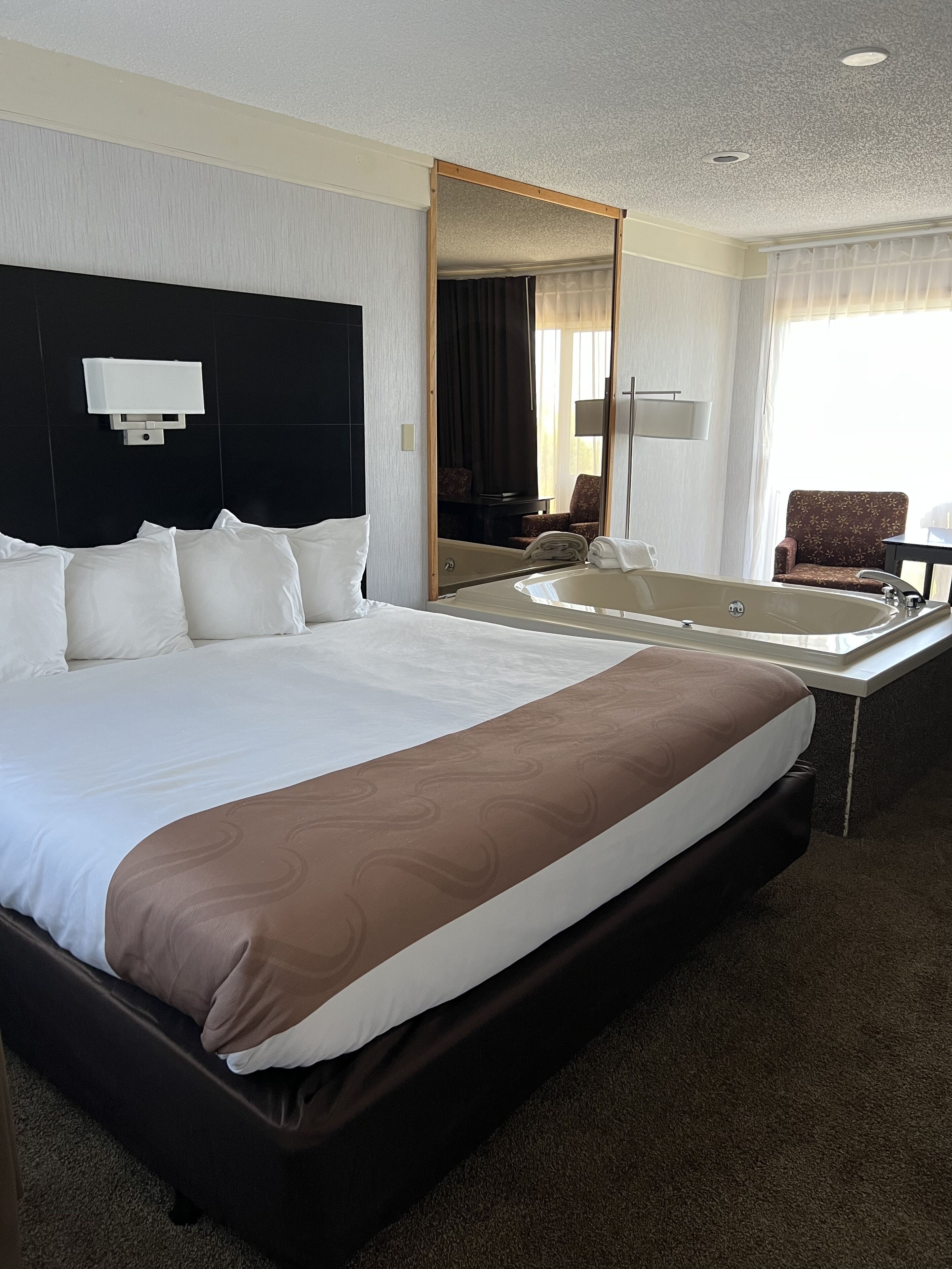 A large bed with a headboard, a bedside table, a mirror, a chair, and a view of the outdoors.