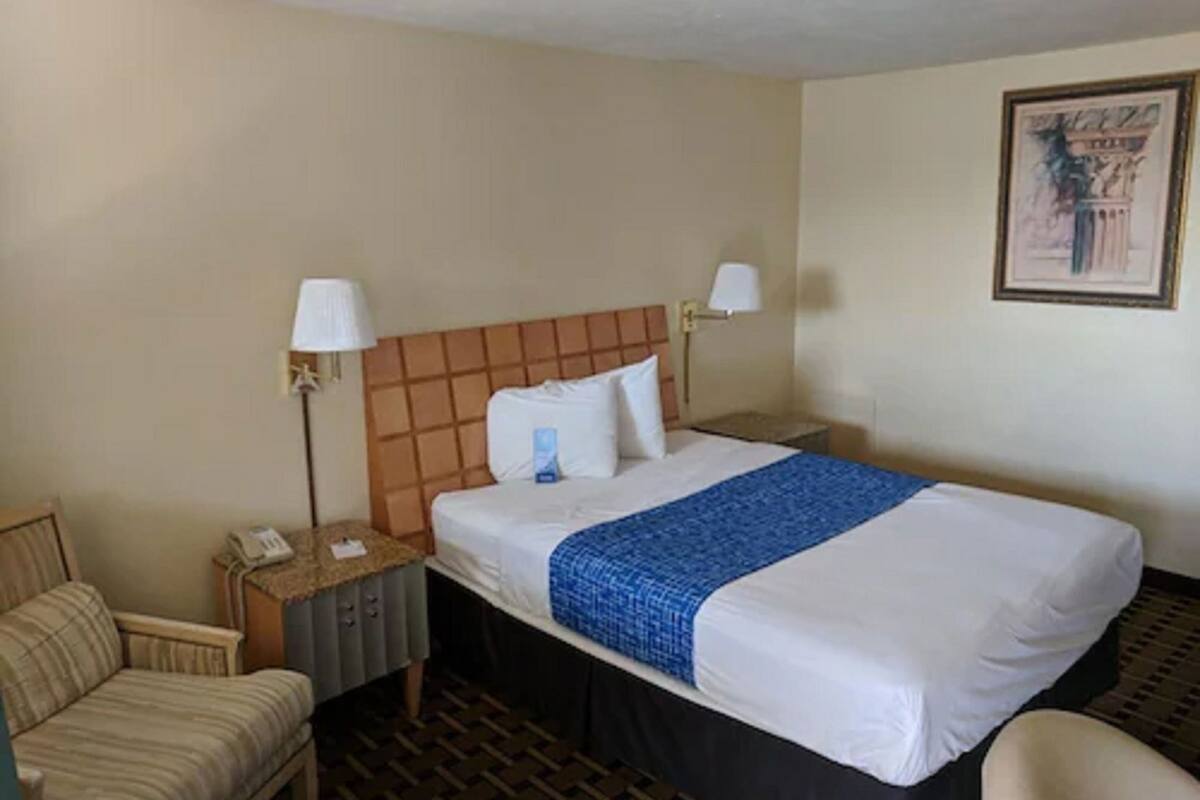 Una habitación de hotel con cama, dos lámparas de noche, mesita de noche, teléfono y una silla.