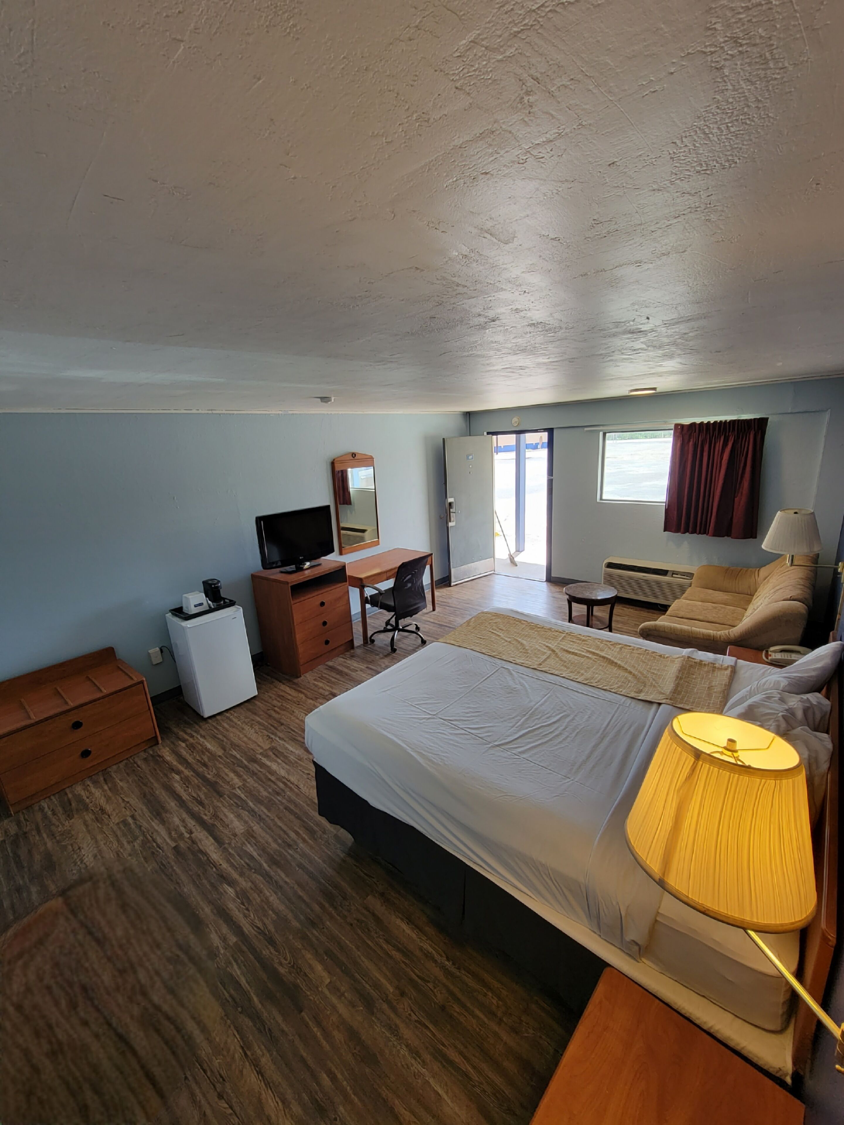 Habitación de hotel con cama, escritorio con silla, una mesita, sofá, televisor, espejo y ventana con cortinas.